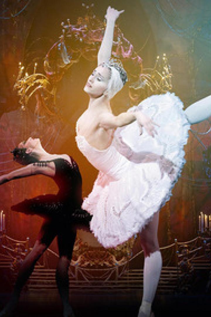 Zwei Ballerinen, eine in Weiß und eine in Schwarz, tanzen auf einer Bühne vor einem dunklen Hintergrund.