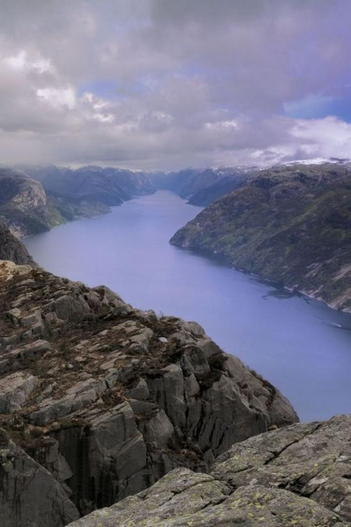 Blick von einem felsigen Gipfel auf einen Fjord, umgeben von Bergen unter einem bewölkten Himmel.