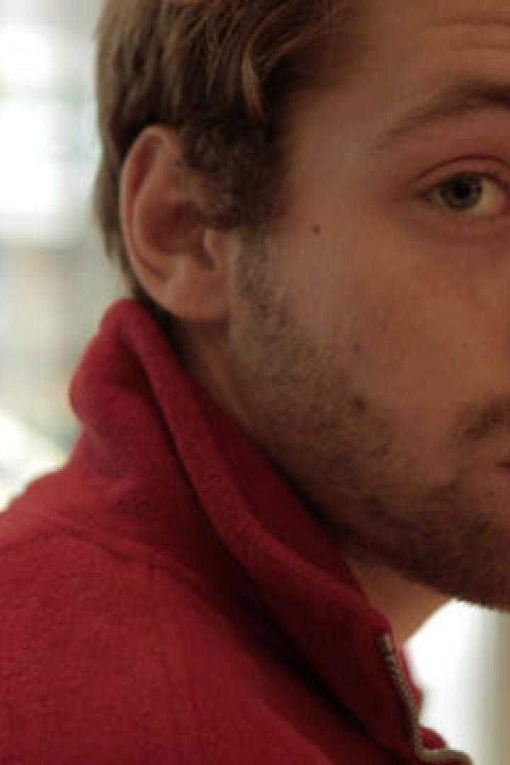 Ein junger Mann mit Bart und roter Jacke blickt über seine Schulter.