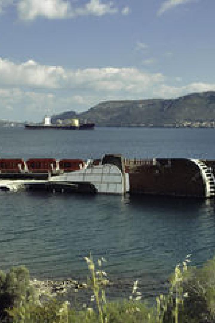 Ein gekentertes Schiff liegt im flachen Wasser vor einer bergigen Küstenlandschaft.