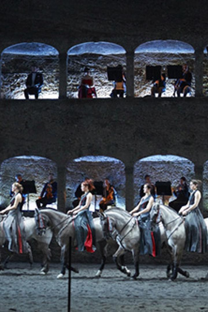 Eine Gruppe von Reiterinnen auf Schimmeln, begleitet von einem Orchester in einer Arena.