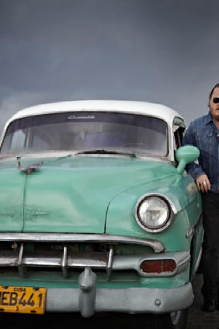 Ein Mann lehnt an einem türkisfarbenen Oldtimer vor einer Skyline unter einem bewölkten Himmel.