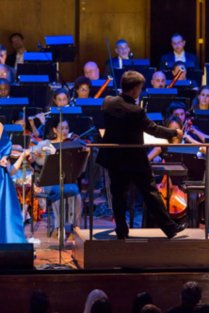Ein Orchester spielt mit einem Sänger, einer Sängerin und einem Violinisten auf einer Bühne.