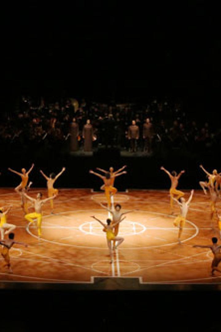 Eine Bühnenaufführung mit Tänzern in einem Kreis und einem Chor im Hintergrund.