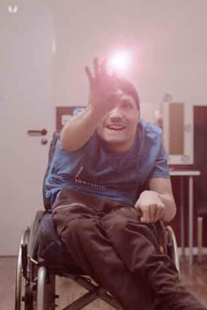 Ein junger Mann im Rollstuhl winkt in einem hellen Raum.