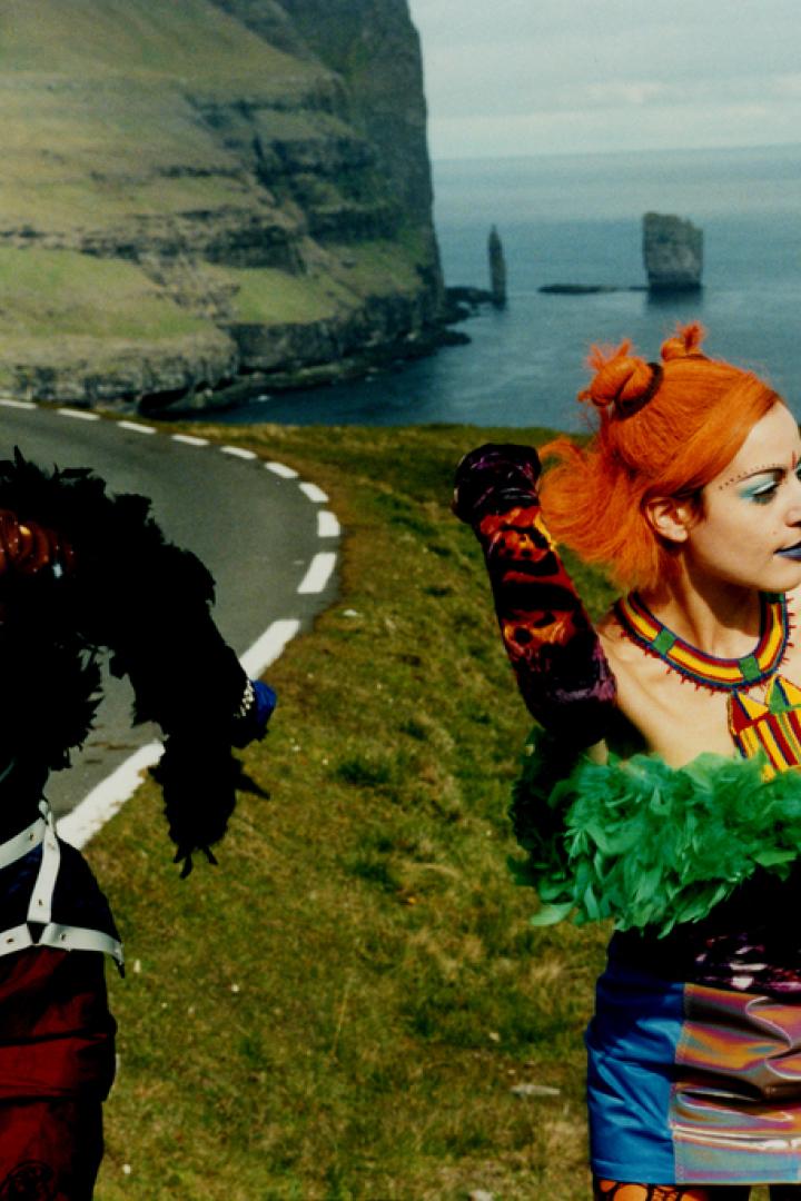 Zwei Frauen in auffälligen Kostümen posieren auf einer Straße vor einer Küstenlandschaft.