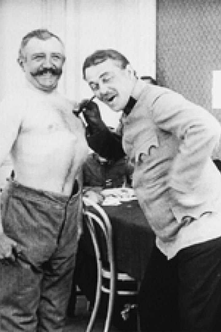 Ein Mann mit freiem Oberkörper und ein uniformierter Mann stehen nebeneinander.