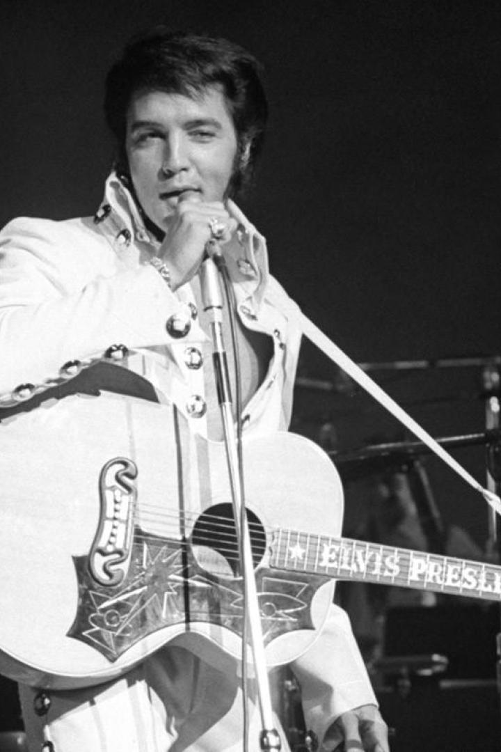 Elvis Presley tritt in einem weißen Anzug mit Gitarre auf einer Bühne auf.