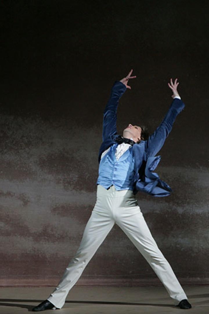 Ein Tänzer in einem blauen Anzug steht mit erhobenen Armen auf einer Bühne.