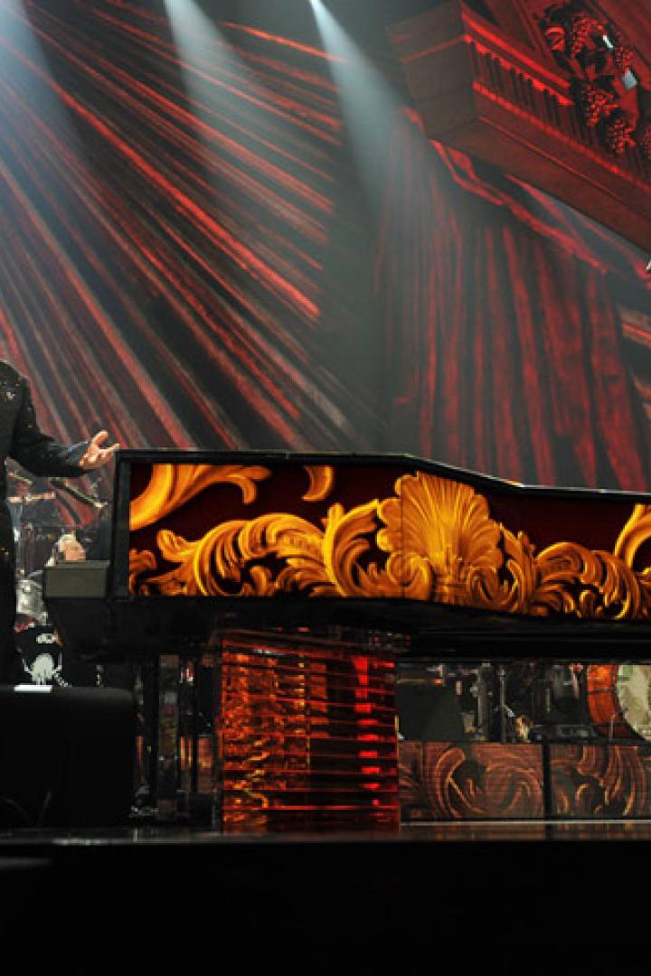 Elton John steht auf einer Bühne mit einem verzierten Klavier und singt.