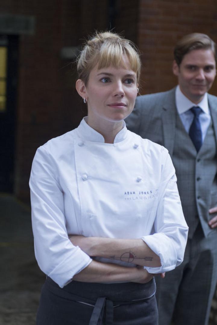 Sienna Miller als Köchin mit verschränkten Armen, Daniel Brühl im Hintergrund.