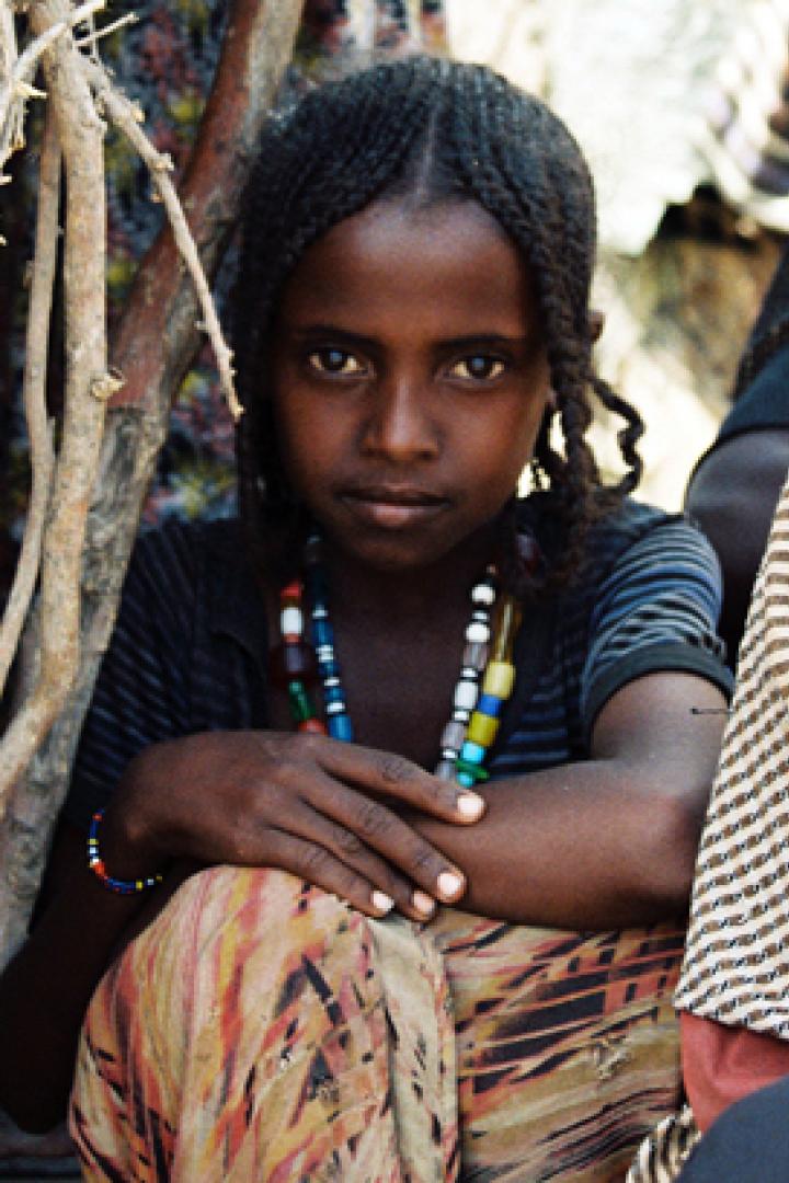 Ein junges Mädchen mit geflochtenen Zöpfen und einer bunten Halskette sitzt im Freien.