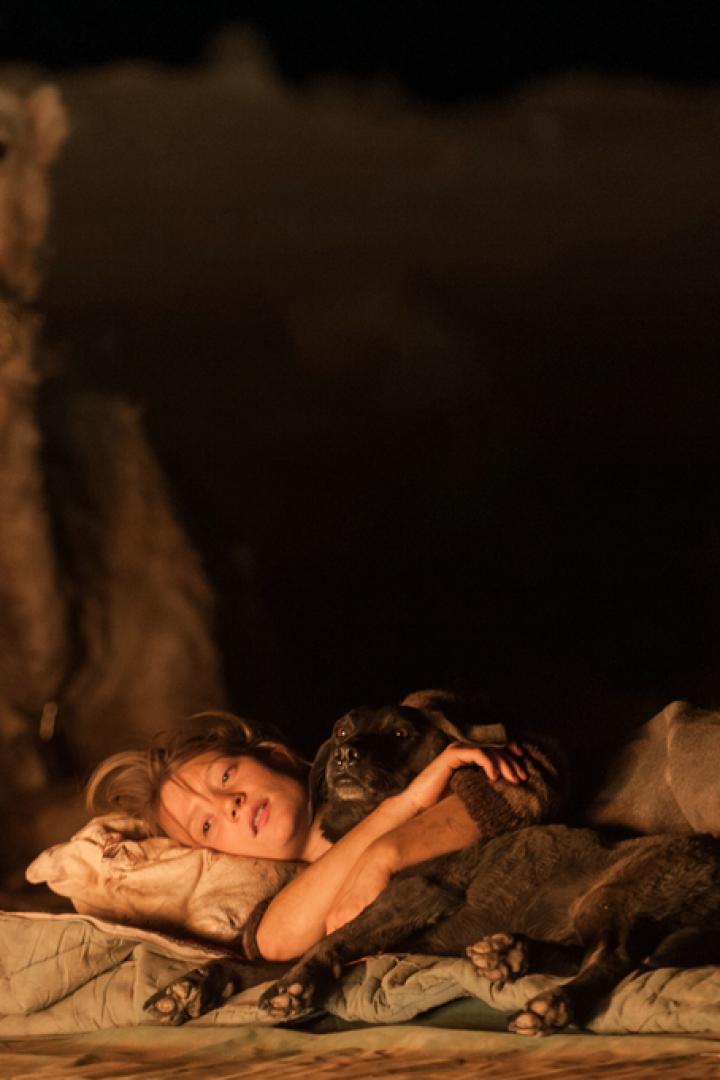 Eine Frau liegt mit einem Hund in der Wüste, im Hintergrund ein Kamel.
