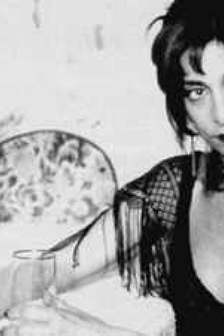 Schwarzweißporträt von Lydia Lunch mit einem Glas und einer Flasche auf dem Tisch.