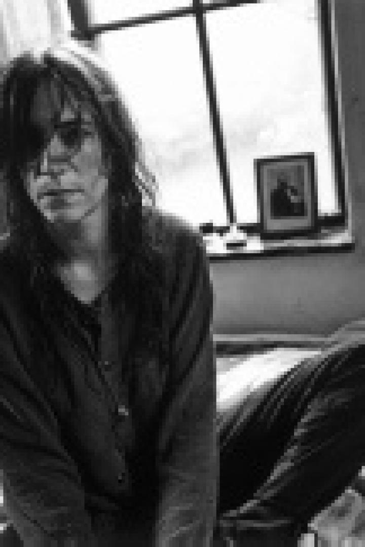Schwarzweißporträt von Patti Smith, die auf einem Bett vor einem Fenster sitzt.