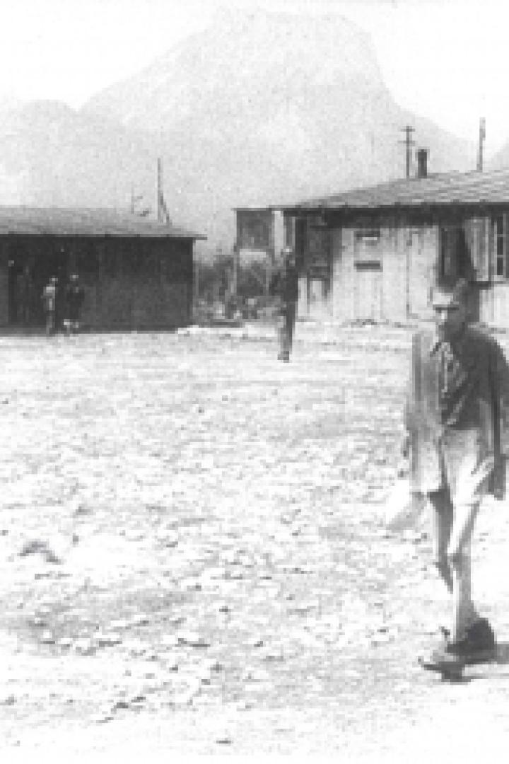 Ein Mann in Shorts und Hemd geht über einen Platz mit Baracken im Hintergrund.