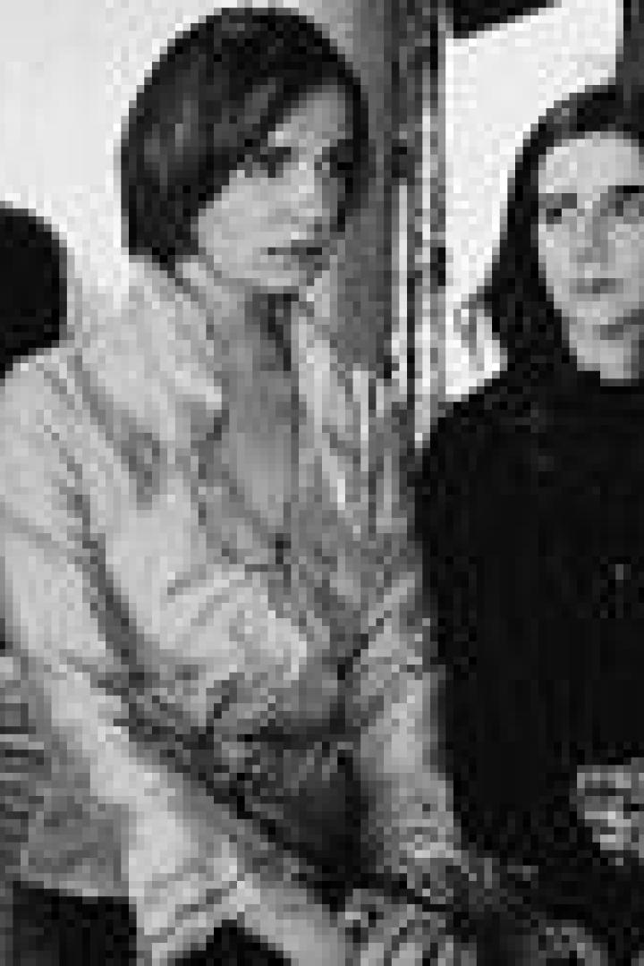 Zwei Frauen sitzen in einem Schlafzimmer, die eine trägt eine Bluse, die andere einen Pullover.