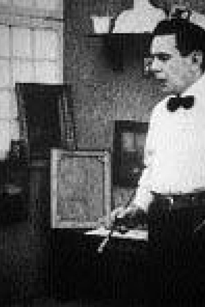 Ein Mann mit Fliege steht in einem Atelier und hält Pinsel und Palette.