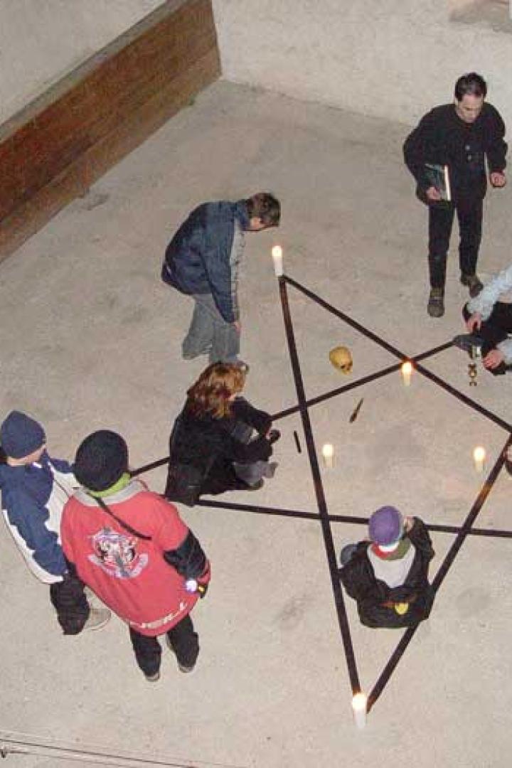 Eine Gruppe von Menschen steht um einen Pentagramm-Altar mit Kerzen und einem Schädel.