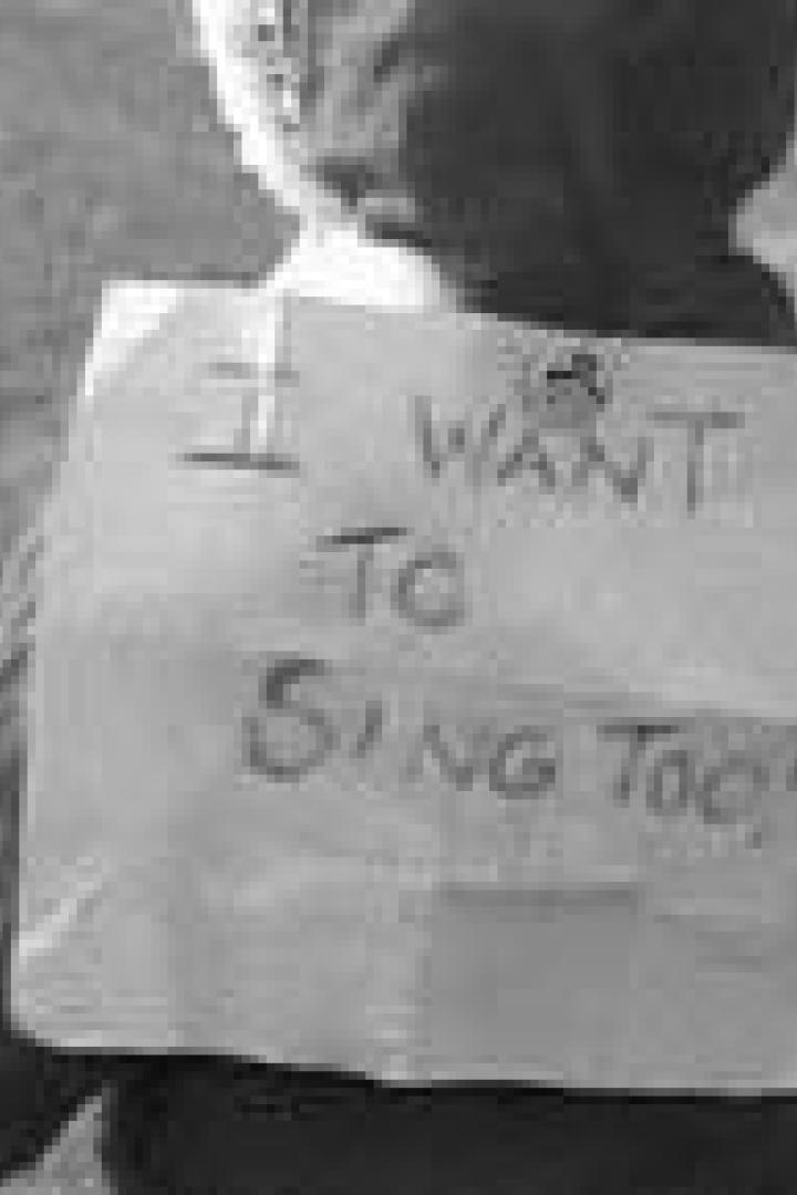 Ein Kind trägt ein Schild mit der Aufschrift „I WANT TO SING TOO!“.