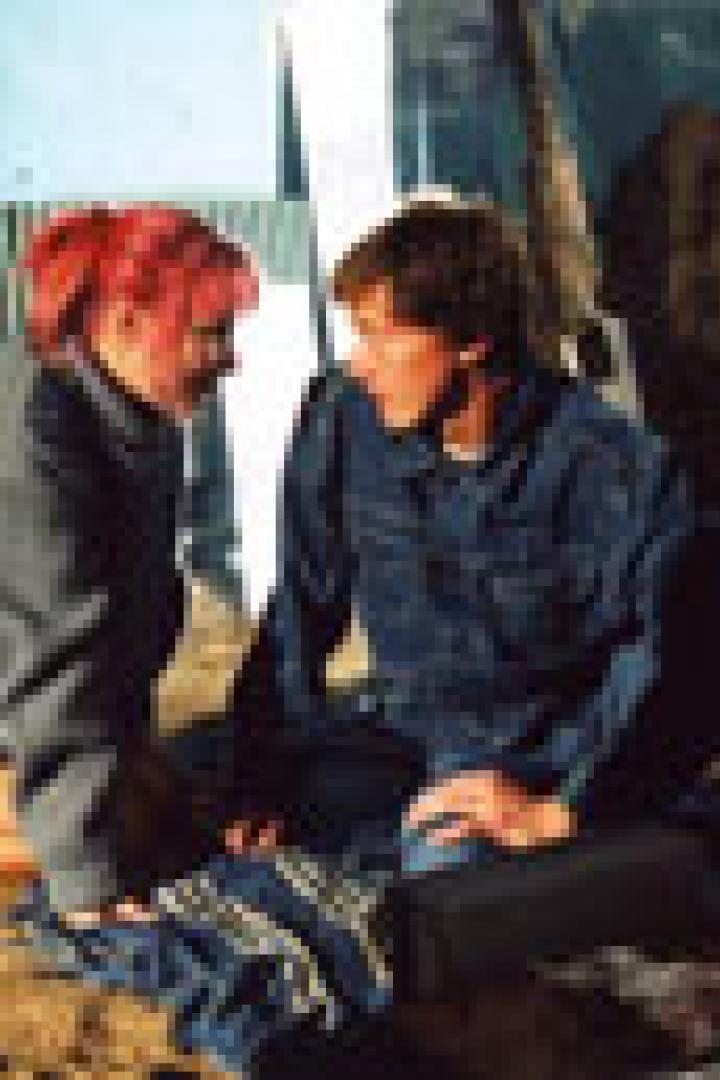 Eine junge Frau mit pinken Haaren und ein junger Mann sitzen auf dem Boden und schauen sich an.
