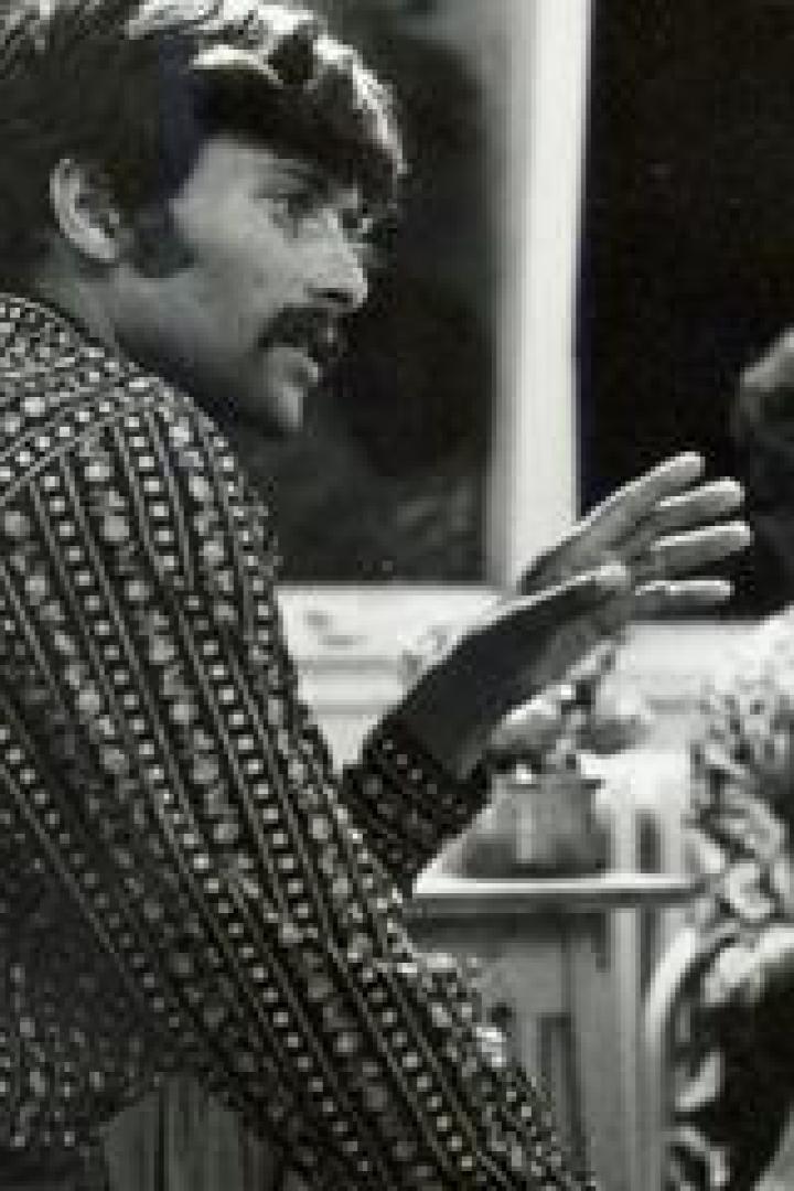 Ein Mann mit Schnurrbart gestikuliert, während ein anderer Mann im Hintergrund sitzt.