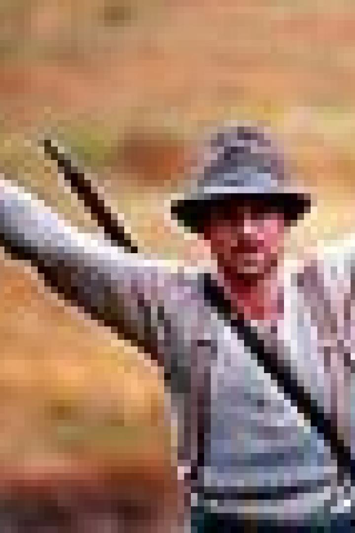 Ein Mann mit Hut und Gewehr steht mit erhobenen Armen in der Natur.