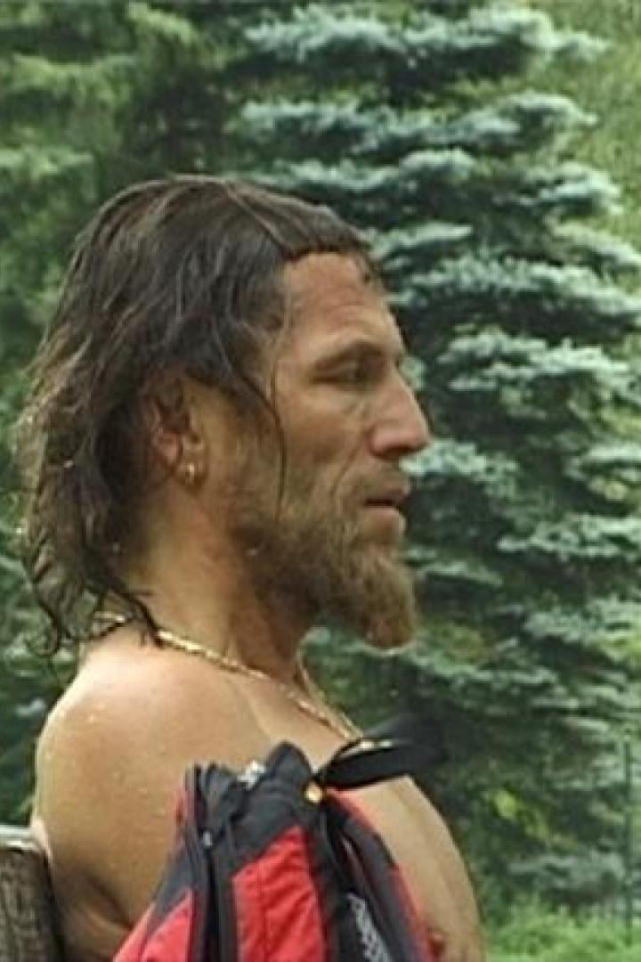 Ein Mann mit Bart und nacktem Oberkörper sitzt auf einer Bank vor Bäumen.