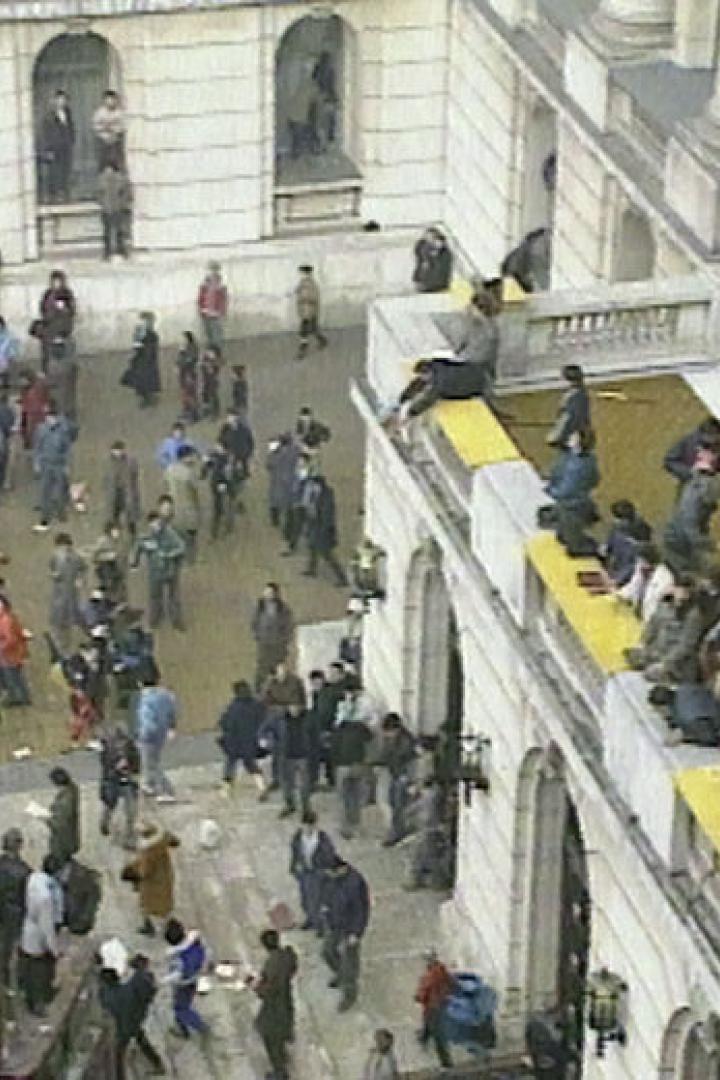 Eine Menschenmenge versammelt sich vor einem Gebäude, einige auf dem Dach, möglicherweise während einer Demonstration.