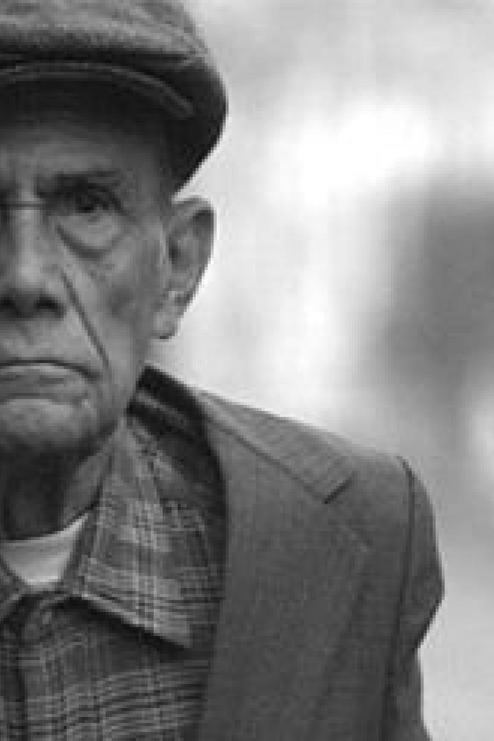 Ein älterer Mann mit einer Schiebermütze und kariertem Hemd blickt ernst in die Kamera.