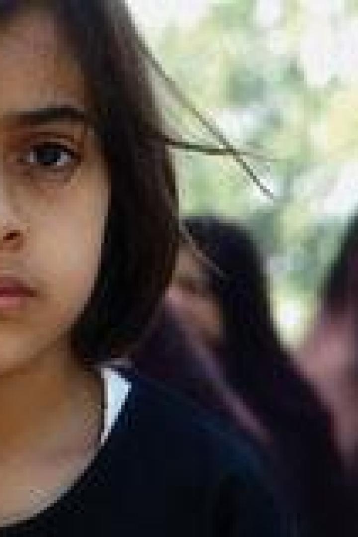 Ein ernstes junges Mädchen steht vor einer Gruppe von Frauen mit Kopftüchern.