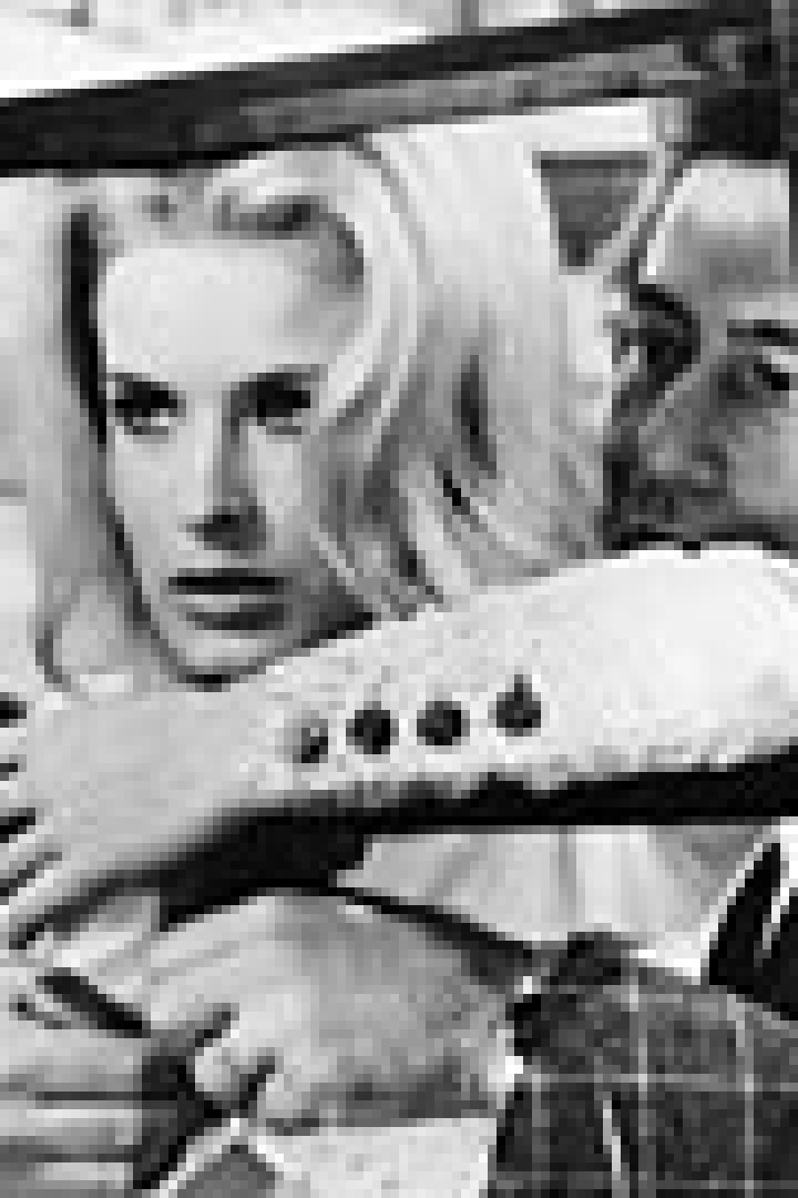 Eine blonde Frau wird von einem Mann umarmt, beide stehen hinter einem dunklen Rahmen.