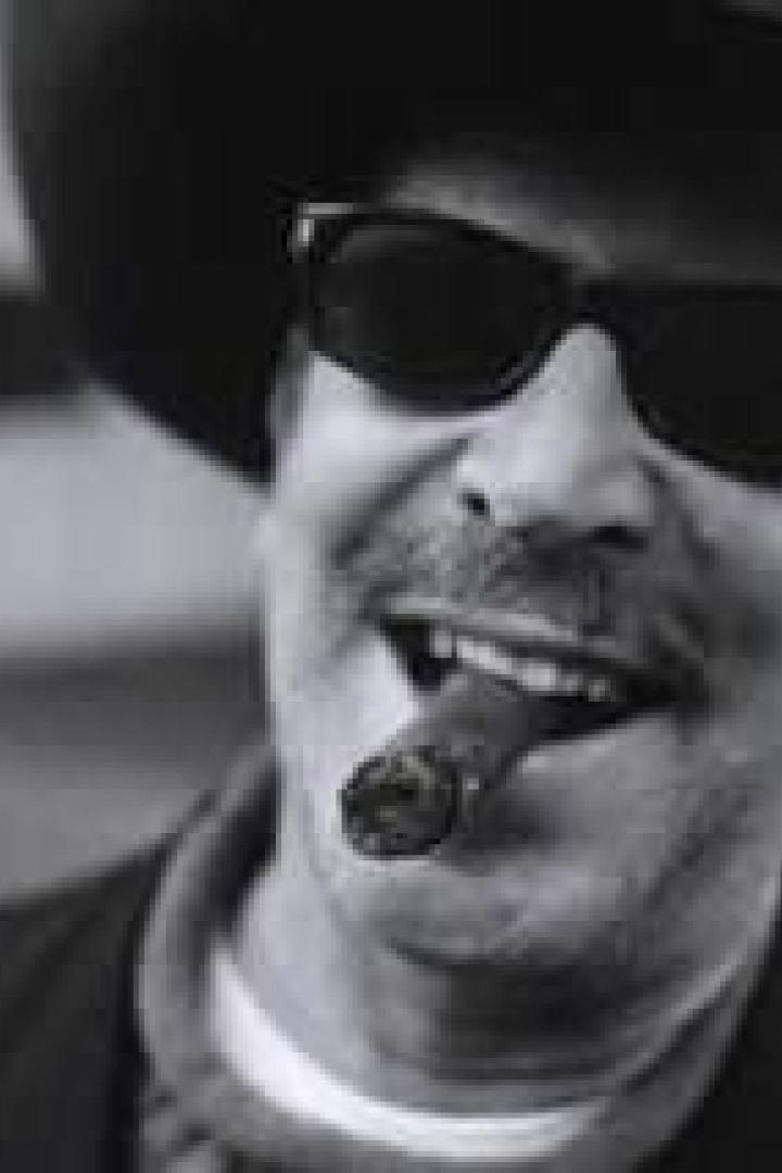 Ein Mann mit Hut, Sonnenbrille und Zigarre lächelt.