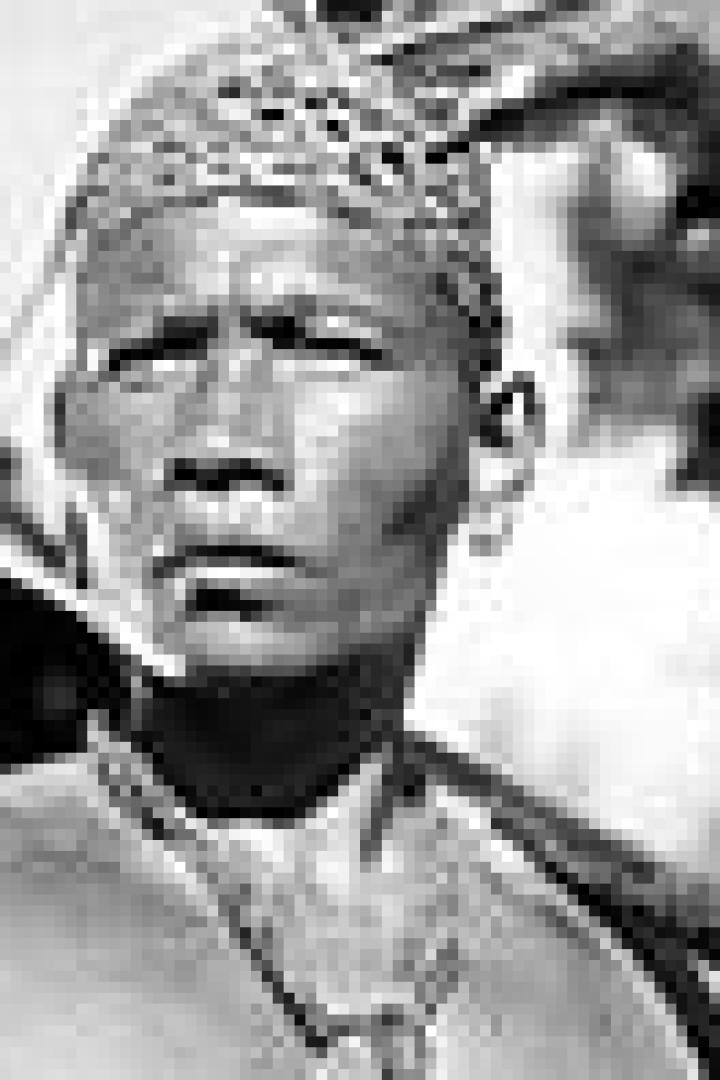 Ein Mann mit Kopfschmuck und traditioneller Halskette trägt einen Gegenstand über der Schulter.