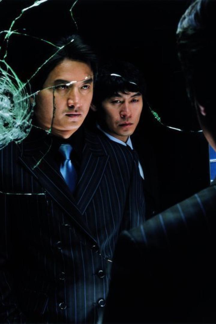 Zwei Männer in Anzügen stehen hinter einem zerbrochenen Spiegel.