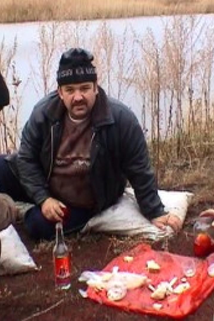 Drei Männer sitzen am Ufer eines Sees und trinken Wodka und essen Snacks.
