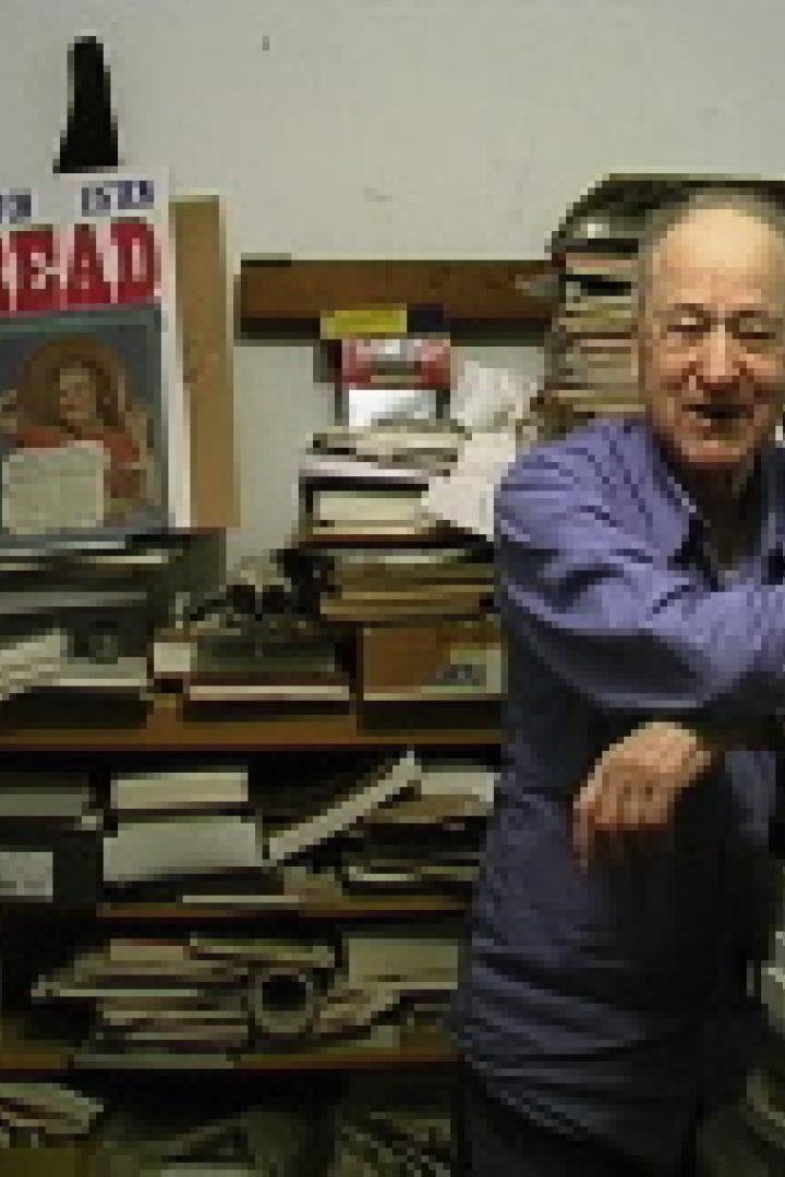 Ein älterer Mann steht vor einem Stapel Kartons in einem mit Büchern und Papieren gefüllten Raum.