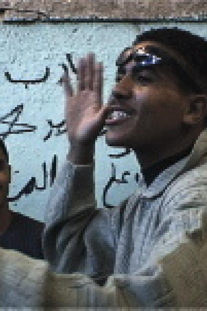 Ein junger Mann gestikuliert vor einer Wand mit arabischer Schrift.