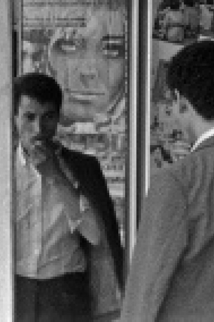 Ein Mann mit Schnurrbart steht vor einem Plakat, während eine andere Person vorbeigeht.