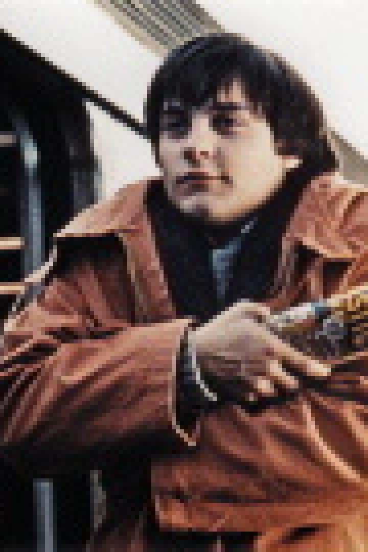 Ein junger Mann in einer orangefarbenen Jacke steht mit verschränkten Armen vor einem Zug.