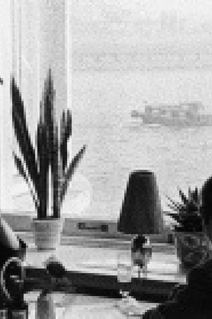 Zwei Personen sitzen an einem Tisch vor einem Fenster mit Blick auf ein Gewässer und ein Schiff.