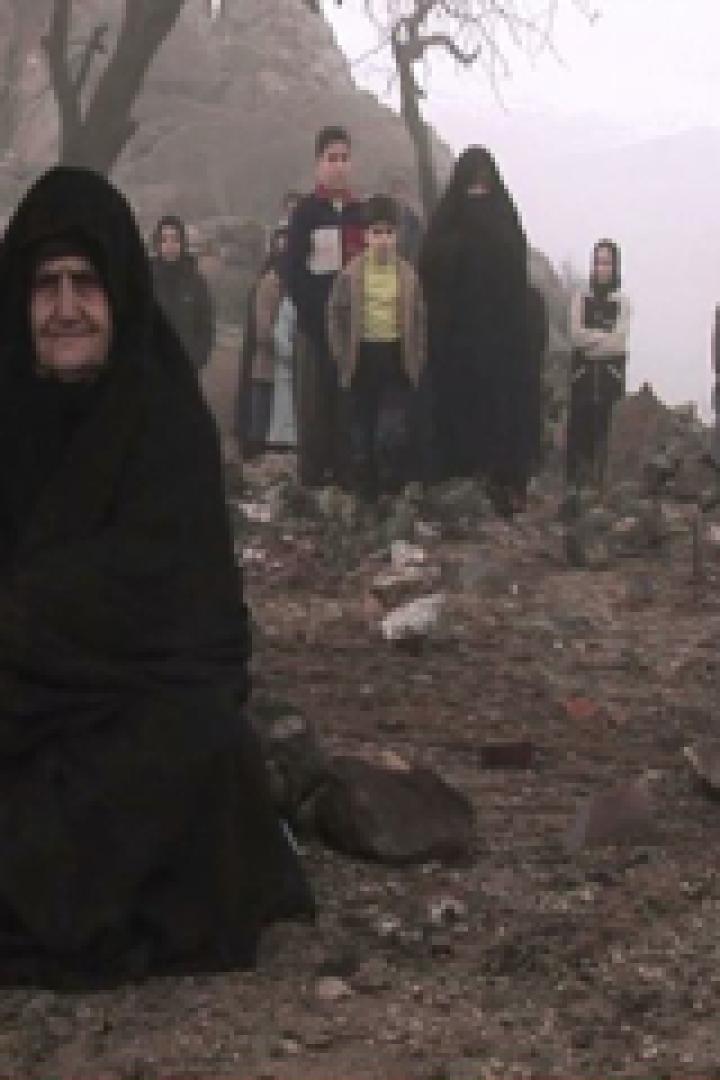 Eine ältere Frau in traditioneller Kleidung sitzt vor einer Gruppe von Menschen auf einem Friedhof.