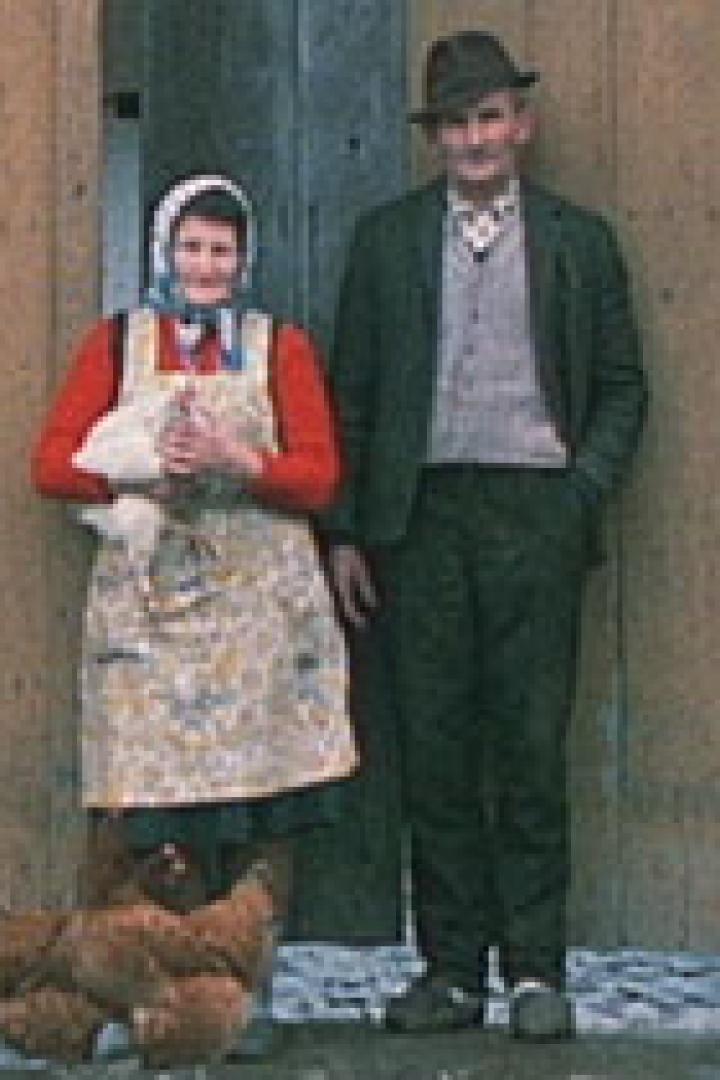 Ein älteres Paar steht vor einer Holzwand, die Frau hält ein Huhn, während Hühner um sie herum laufen.