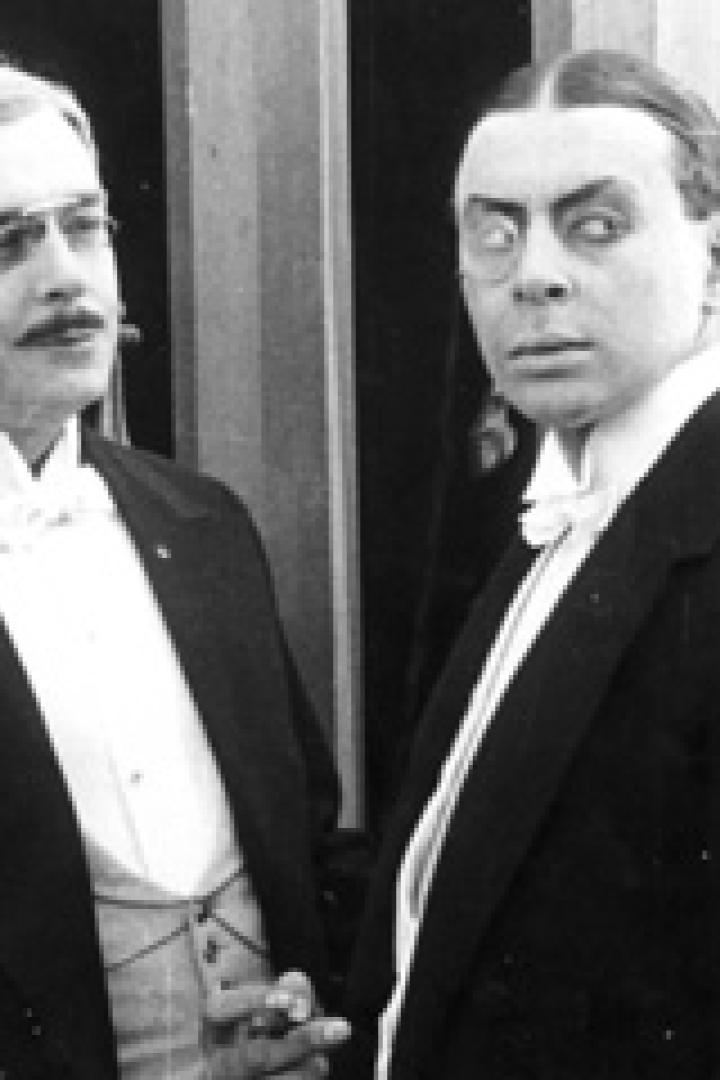 Zwei Männer in Anzügen, einer mit Brille und Schnurrbart, der andere mit starrem Blick.