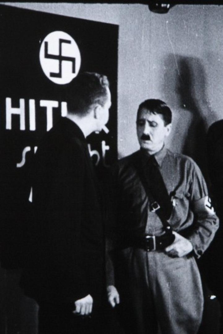 Eine Schwarzweißaufnahme von Adolf Hitler mit zwei Männern vor einem Hakenkreuz-Schild.