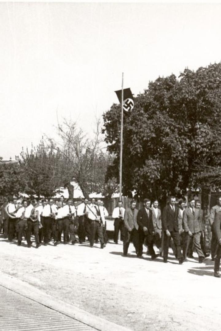 Ein Schwarzweißfoto einer Parade mit Hakenkreuzflagge und Bannern in einer Stadt.