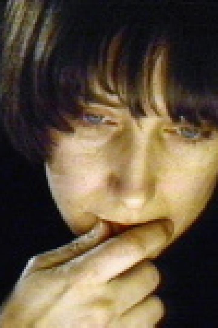 Eine Frau mit dunklen Haaren und hellen Augen blickt nach unten und berührt ihre Lippen.