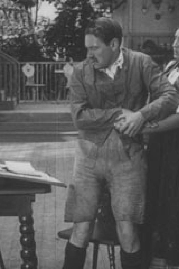 Eine Frau hält einen Mann fest, der sich vor Schmerzen krümmt, während ein Tisch mit Papieren im Vordergrund steht.