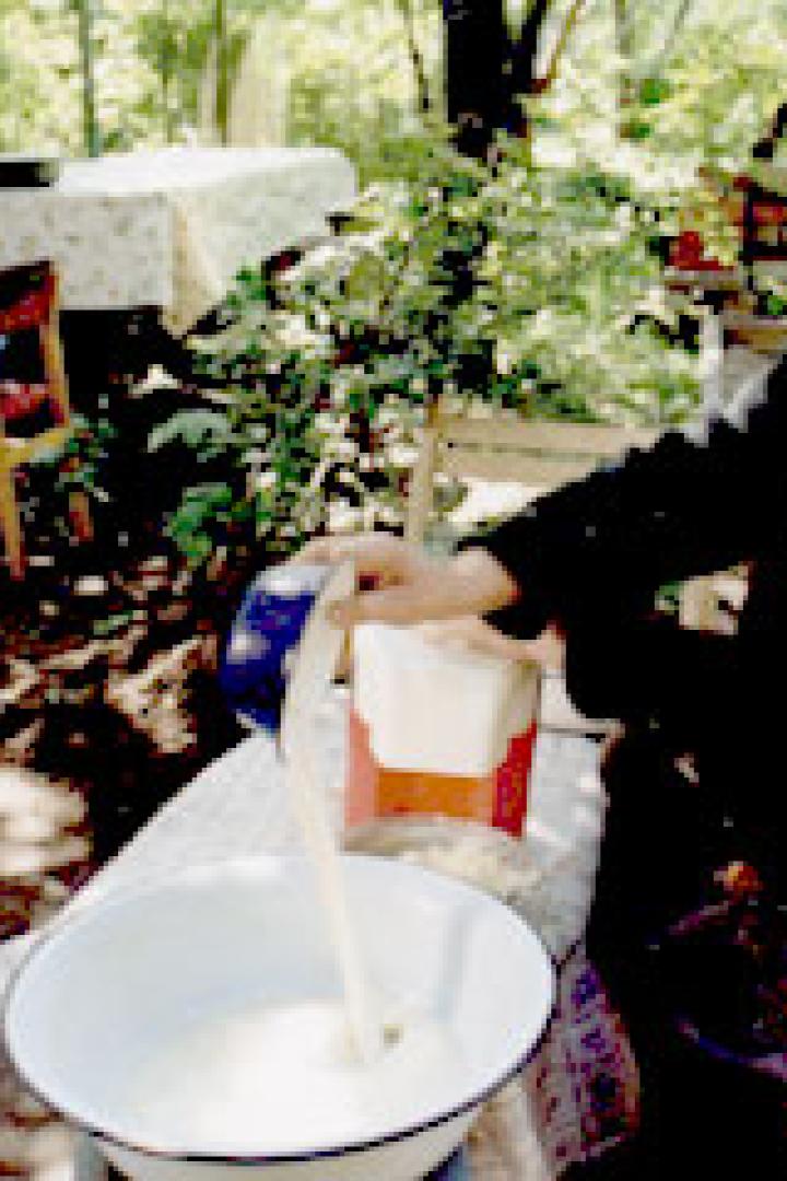 Ein älterer Mann gießt Milch in eine Schüssel im Freien, während eine Frau im Hintergrund steht.