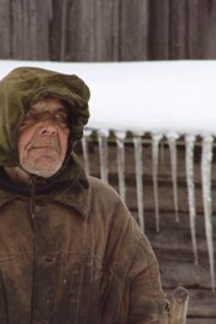 Ein älterer Mann mit wettergegerbtem Gesicht und Kapuze steht vor einem schneebedeckten Holzgebäude mit Eiszapfen.